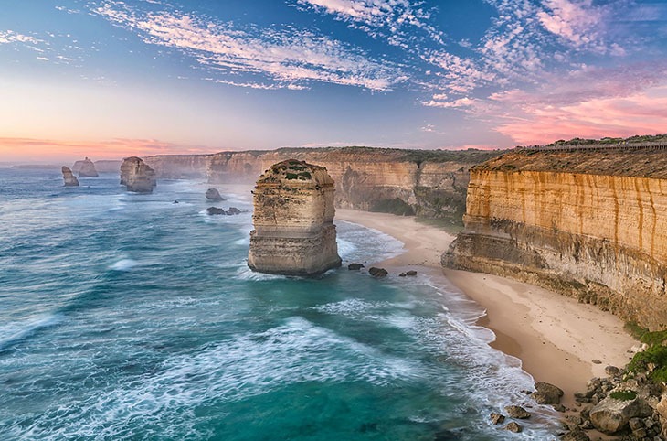 A scenic coastal view with rocky cliffs, ocean waves, and sea stacks under a colorful sunset sky.